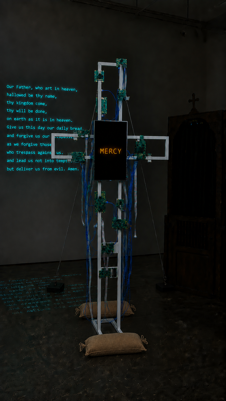 A Digital Prayer: freestanding cross with portrait screen displaying MERCY in amber, PCB panels and ribbon cables across the frame, prayer projected on gallery wall behind